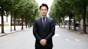 並木道の真ん中で姿勢よく正面を向いて立っている営業マン風の清潔感のある男性