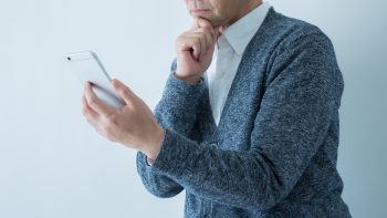 シニア男性がスマートフォンの画面をながめながらもう片方の手を顎に当てて悩んでいる様子